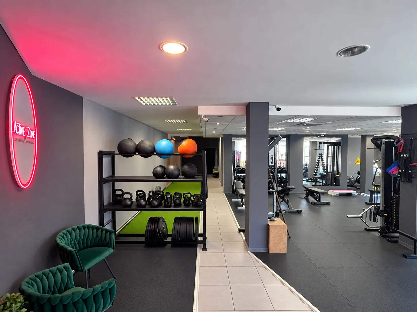 Regał z piłkami lekarskimi, kettlebellami i talerzami przy torze ze sztuczną trawą, obok podświetlane logo studia